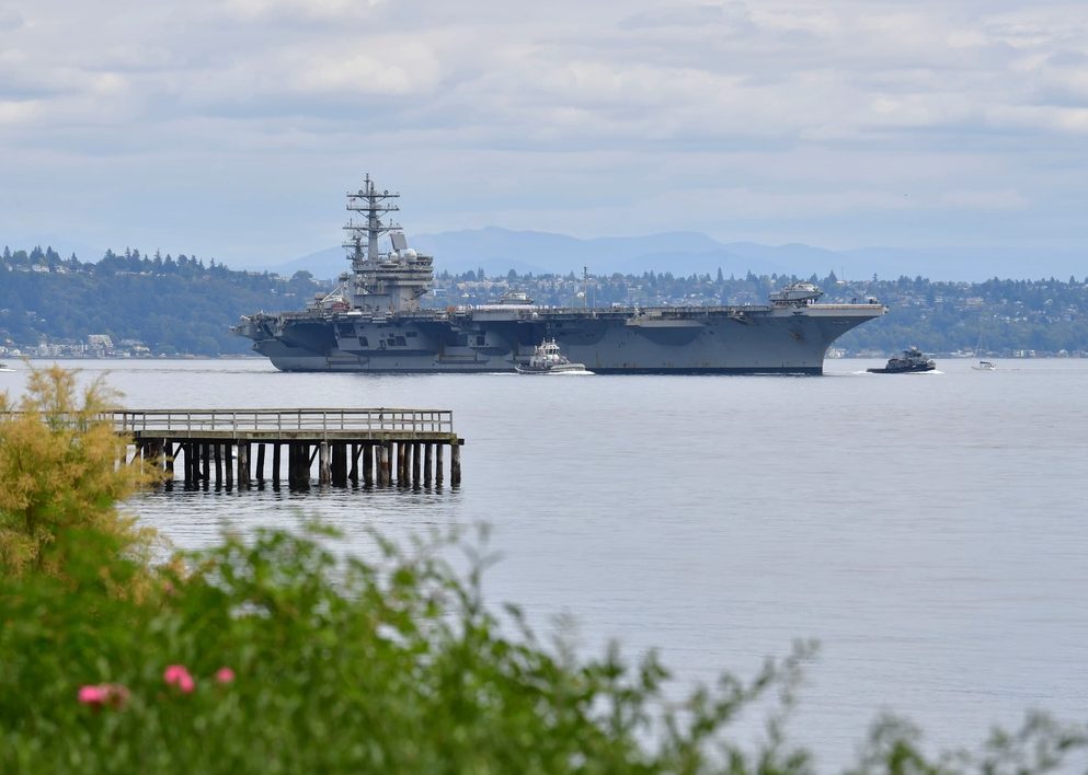 A large aircraft carrier sailing in a calm bay with a wooden pier in the foreground.
